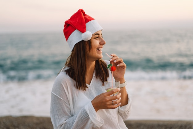 Young beautiful woman in a santa hat with a glass of cocktail in her hands walk on the beach