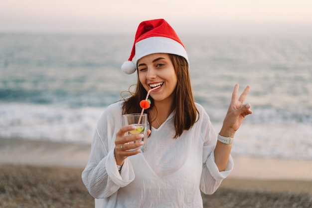 Photo young beautiful woman in a santa hat with a glass of cocktail in her hands walk on the beach