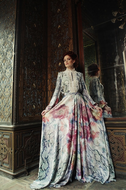 Young beautiful woman standing in the palace room with mirror.