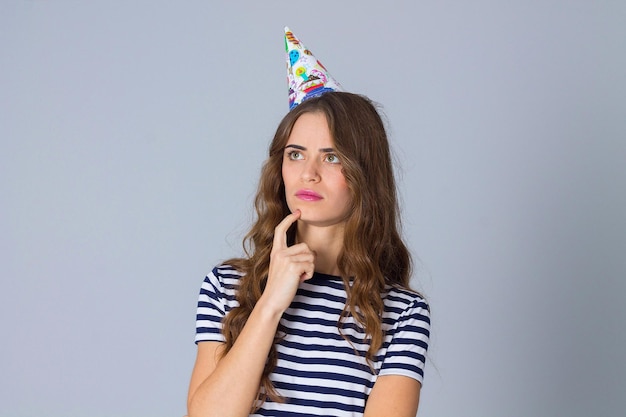 Young beautiful woman in stripped T-shirt and celebration cap thinking on grey background in studio