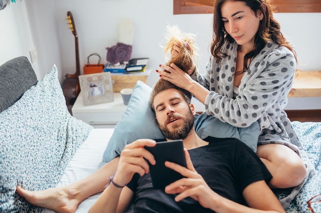 Young beautiful woman stroking hair of bearded man while he is using tablet