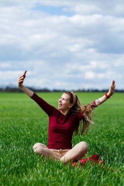 Young beautiful woman student with a phone in her hands sitting on the grass. Girl takes selfies and takes selfie pictures. She smiles and enjoys a warm day. Concept photo on smartphone