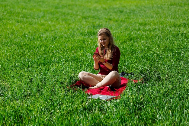 Young beautiful woman student with a phone in her hands sitting on the grass. Girl takes selfies and takes selfie pictures. She smiles and enjoys a warm day. Concept photo on smartphone