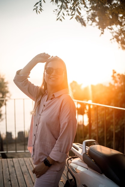 A young beautiful woman in summer clothes stands on the background of an orange sunset near the city moped selective focus