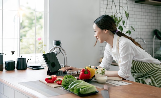 Young beautiful woman using laptop computer searching and learning for cooking healthy food from fresh vegetables and fruits in kitchen room