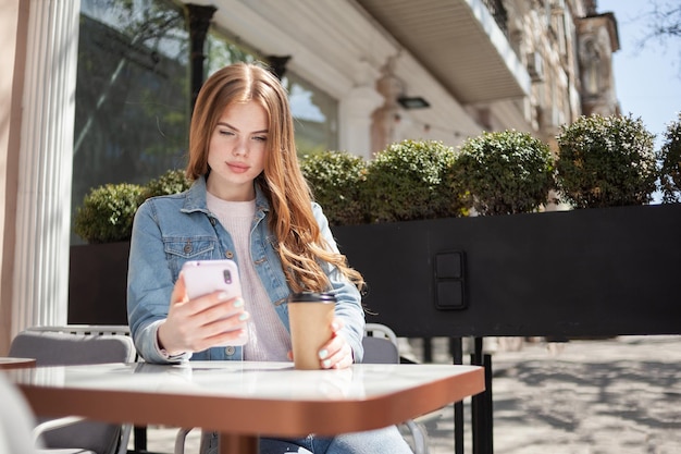 Young beautiful woman using smartphone and drinking coffee while sitting at a table in outdoor cafe