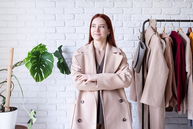 Young beautiful woman wearing beige pastel coat standing near clothes rack wardrobe change