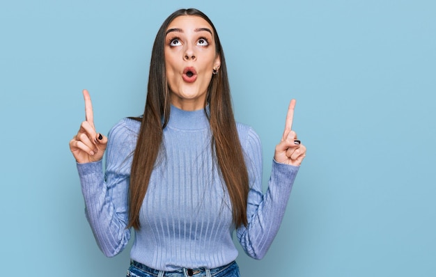 Young beautiful woman wearing casual winter sweater amazed and surprised looking up and pointing with fingers and raised arms