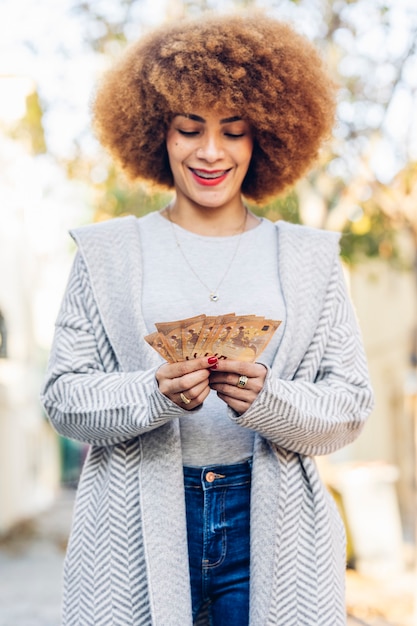 Young and beautiful woman with afro hair smiling happy. Standing with smile on face holding euro bills on city street