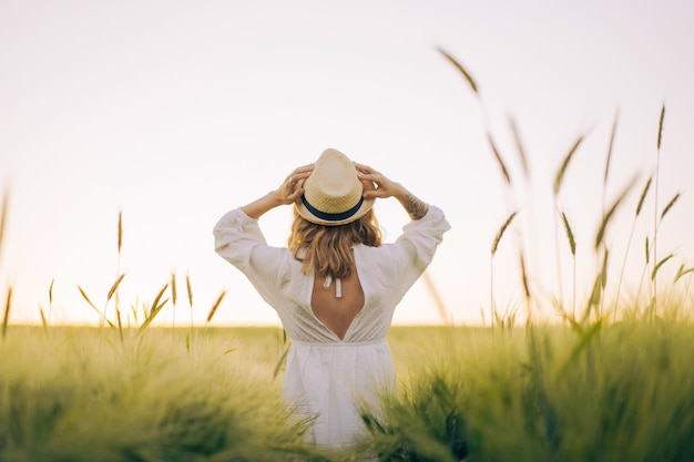 young beautiful woman with blond long hair in a white dress in a straw hat collects flowers on a wheat field. Flying hair in the sun, summer. Time for dreamers, golden sunset.