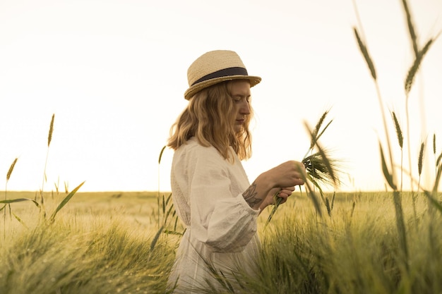 young beautiful woman with blond long hair in a white dress in a straw hat collects flowers on a wheat field. Flying hair in the sun, summer. Time for dreamers, golden sunset.