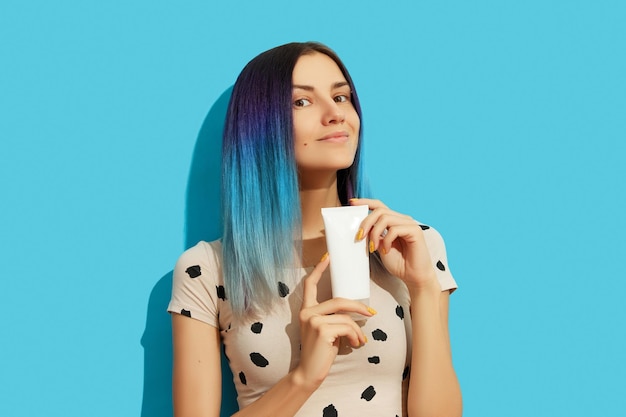 Young beautiful woman with blue hair holding white cosmetic bottle on bright background