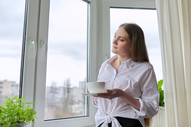 Young beautiful woman with cup of tea at home near window