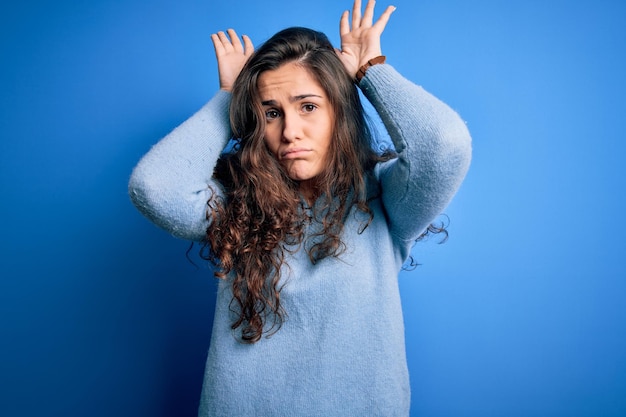 Young beautiful woman with curly hair wearing blue casual sweater over isolated background Doing bunny ears gesture with hands palms looking cynical and skeptical Easter rabbit concept