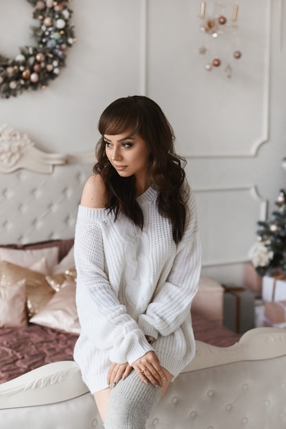 Young beautiful woman with dark hair in fashionable cozy clothes posing in decorated for Christmas bedroom.