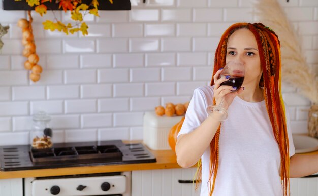 Young beautiful woman with dreadlocks drinking red wine from glass in kitchen Adult female enjoying leisure weekends holidays