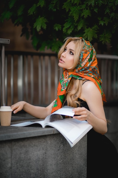 Young beautiful woman with headscarf on head is reading magazine on street there is disposable cup