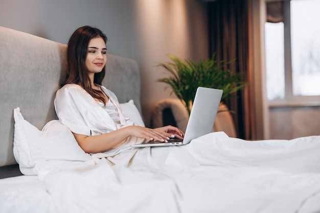 Young beautiful woman with laptop in bed. Brunette model with laptop in silk white pajamas. A woman works at a computer in her bedroom at home.