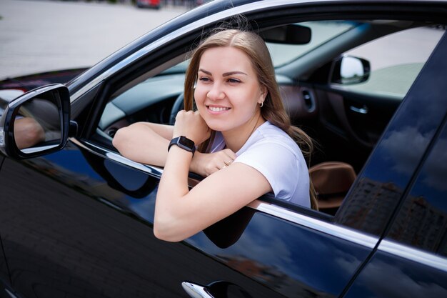 Young beautiful woman with long hair sits in a black car at a parking lot. Pretty girl in casual clothes. Car trip