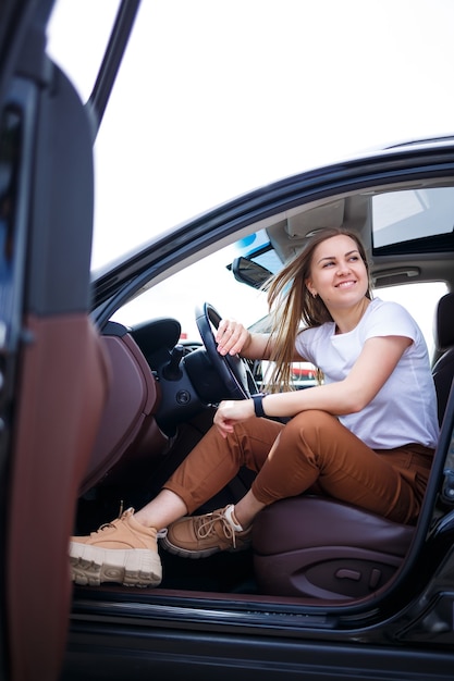 Young beautiful woman with long hair sits in a black car at a parking lot. Pretty girl in casual clothes. Car trip