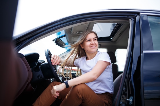 Young beautiful woman with long hair sits in a black car at a parking lot. Pretty girl in casual clothes. Car trip