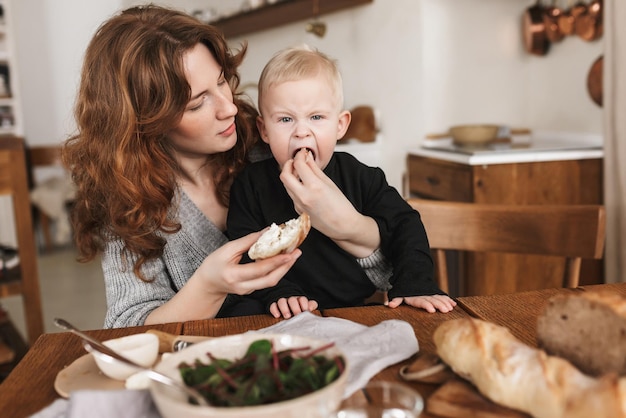 Young beautiful woman with red hair in knitted sweater sitting at the table with food dreamily feeding her little son Mom dreamily spending time with baby boy in cozy kitchen at home