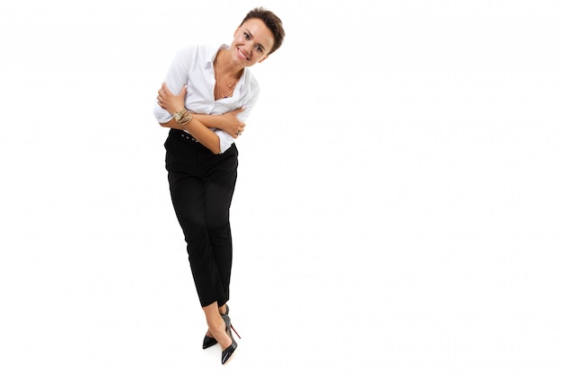 A young beautiful woman with short dark hair, makeup in a white shirt, black pants, with a wristwatch, bracelet and white manicure stands, crossed her legs, leaned slightly and smiles