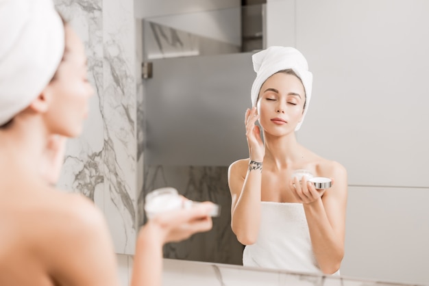 Young beautiful woman wrapped in towels applying cream on her face in the bathroom