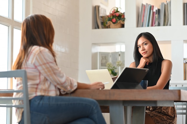 Young beautiful women sitting together in front their computer laptop and tablet at the modern wooden table over living room book shelf