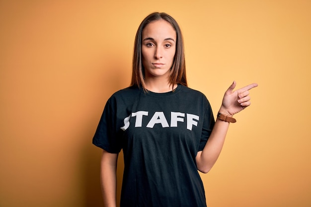 Young beautiful worker woman wearing staff uniform tshirt over isolated yellow background Pointing with hand finger to the side showing advertisement serious and calm face