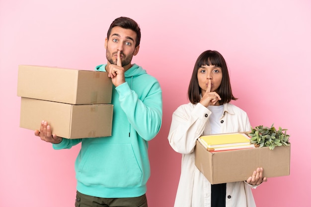 Young beauty couple moving in new home among boxes isolated on pink background showing a sign of silence gesture putting finger in mouth