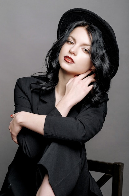 Young beauty fashion model woman in  hat portrait on a gray wall sitting on a chair,