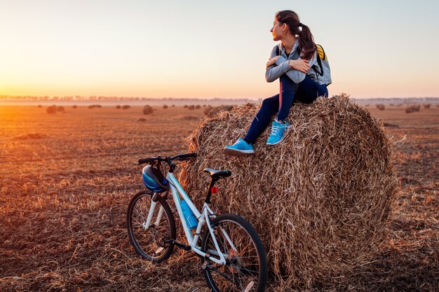 Young bicyclist having rest after a ride in autumn field at sunset. Woman admiring view sitting on haystack. Sport recreation concept