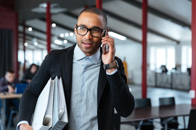 Young bisnessman carrying folders and talking on the phone in the office