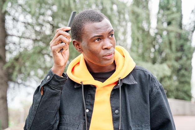 Young black AfricanAmerican man in yellow sweatshirt black jacket is combing his hair in the city