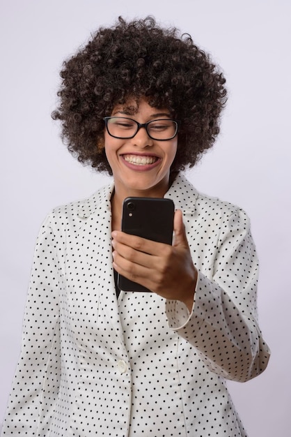 Young black Brazilian woman holding and looking at a cell phone device smiling wearing a white shirt