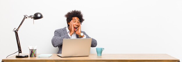 Young black businessman feeling happy, excited and surprised, looking to the side with both hands on face on a desk