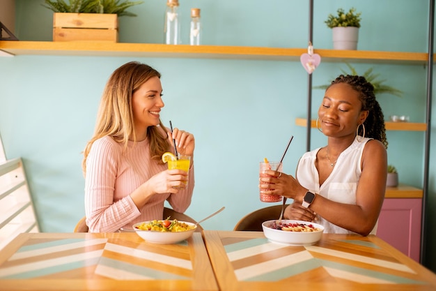 Young black and caucasian woman having good time drinking fresh juices and having healthy breakfast in the cafe