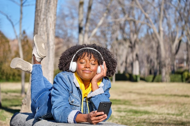 Young black girl lying on a bench listening to music with her eyes closed with her smart phone