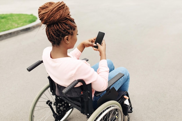 Young black lady in wheelchair using cellphone with mockup for mobile app outdoors