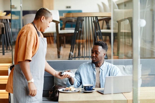 Young black man having lunch in modern cafe sitting at table paying for order with bank card