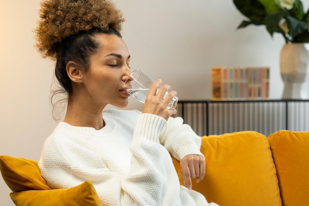 Young black woman drinking a glass of water sitting on the sofa in the living room