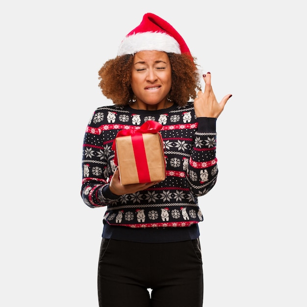 Young black woman holding a gift in christmas day crossing fingers for having luck