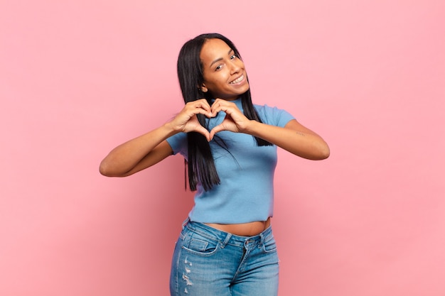 Young black woman smiling and feeling happy, cute, romantic and in love, making heart shape with both hands