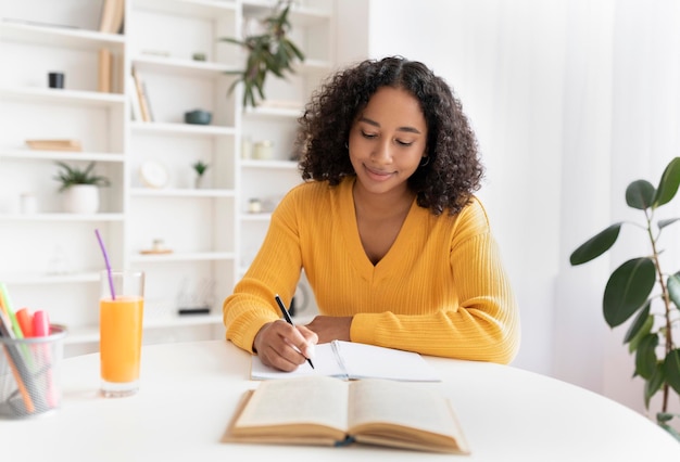 Young black woman writing in notebook getting ready for exam or test sitting at desk indoors