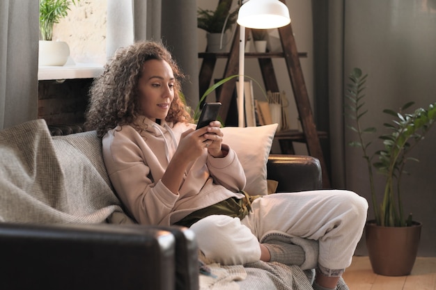 Young blogger sitting on sofa and working online on her mobile phone at home