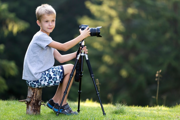 Young blond child boy sitting on tree stump on grassy clearing taking picture with tripod camera.