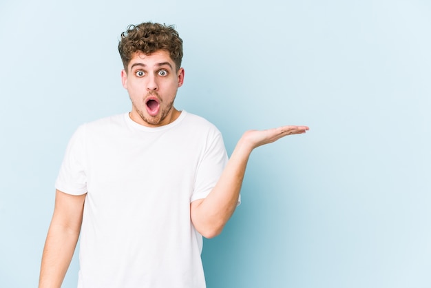 Young blond curly hair caucasian man isolated impressed holding copy space on palm.