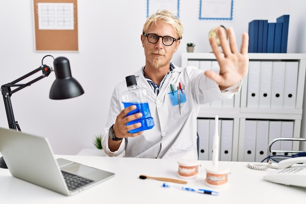 Photo young blond dentist man holding mouthwash at clinic with open hand doing stop sign with serious and confident expression defense gesture