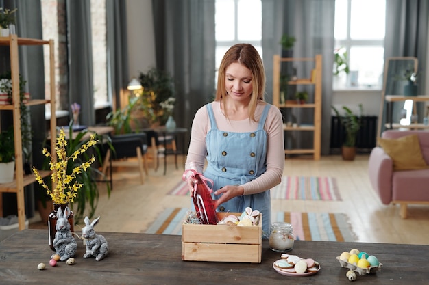 Young blond female putting bottle of fruit drink for children and other snacks into wooden box while standing by table in living-room